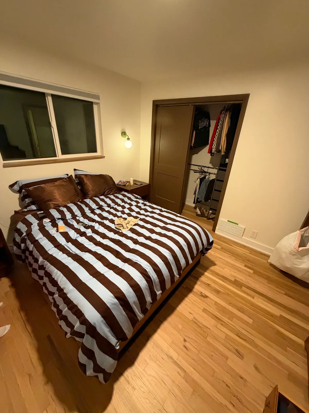 Current bedroom with striped bedding, wood nightstand, green globe sconce and brown sliding closet